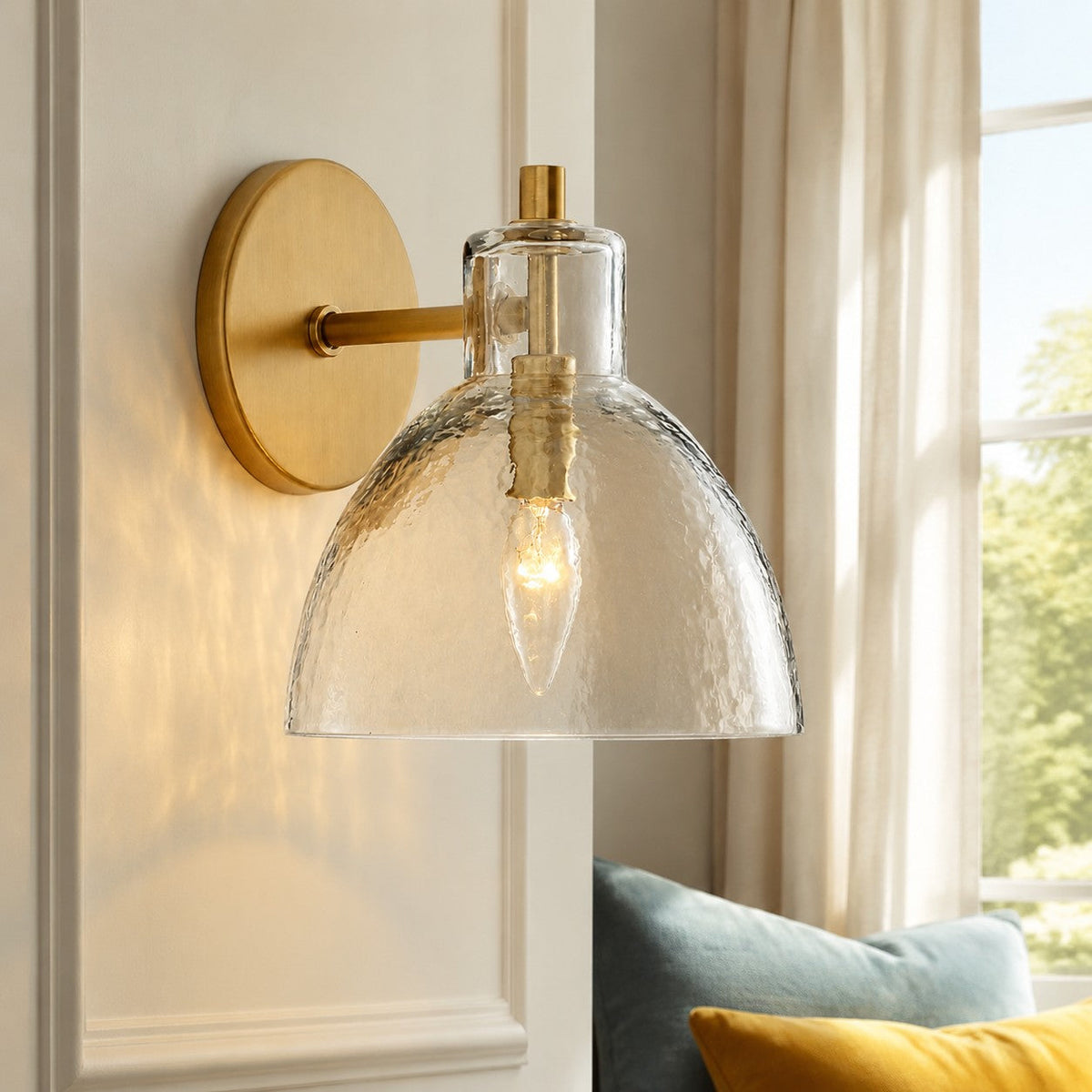 Brass hammered glass wall sconce glowing beside a window in a sunlit room