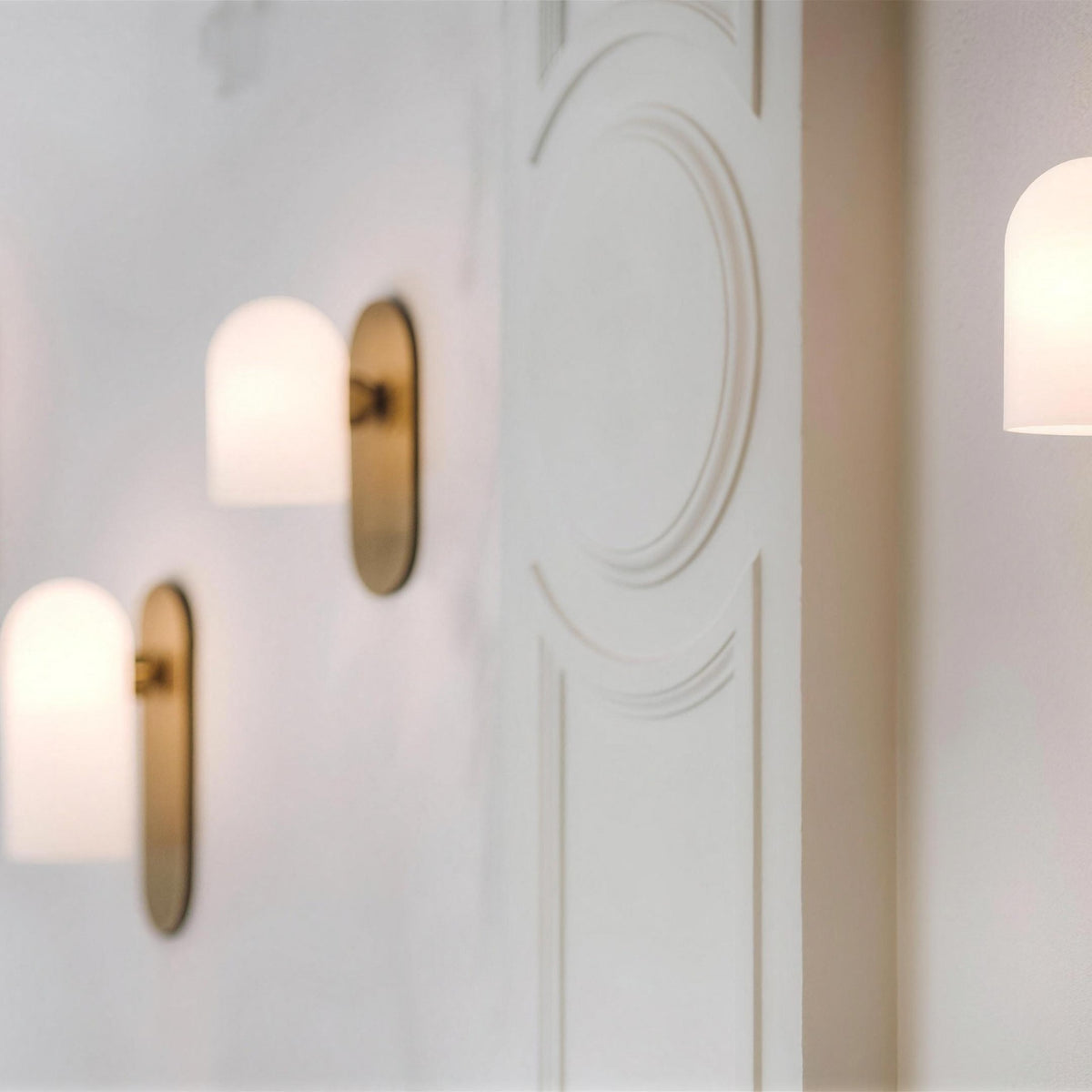 Capsula Opalina brass wall sconces glowing along a paneled white hallway