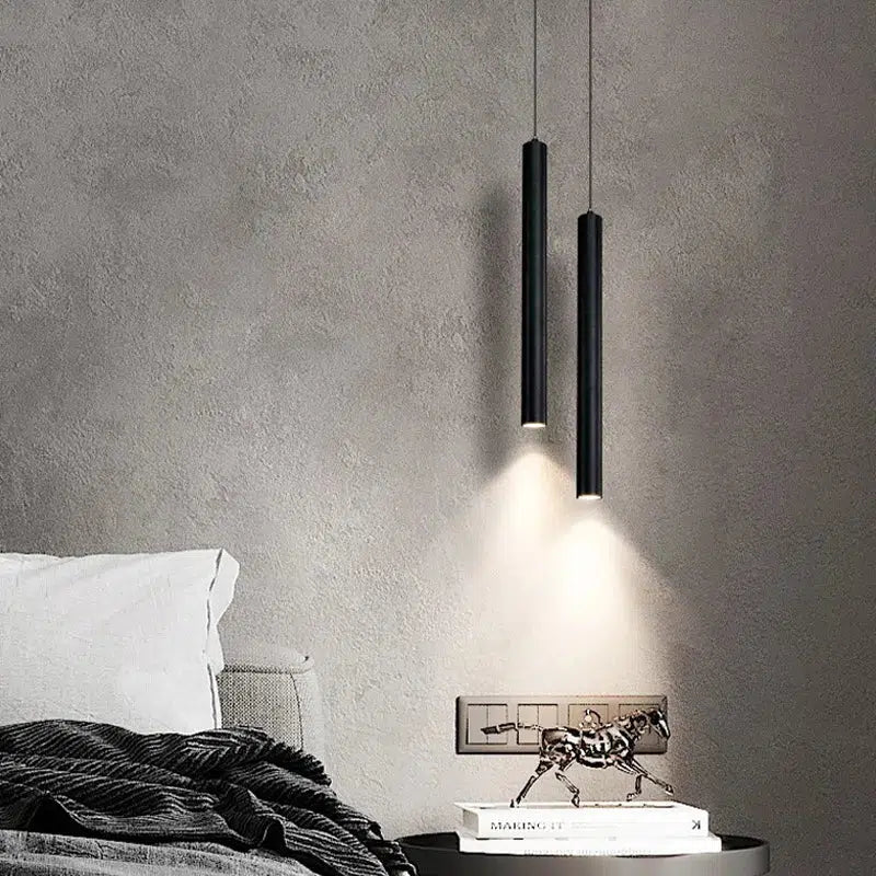 Two black Flauto tube pendants casting warm spotlights beside a bed