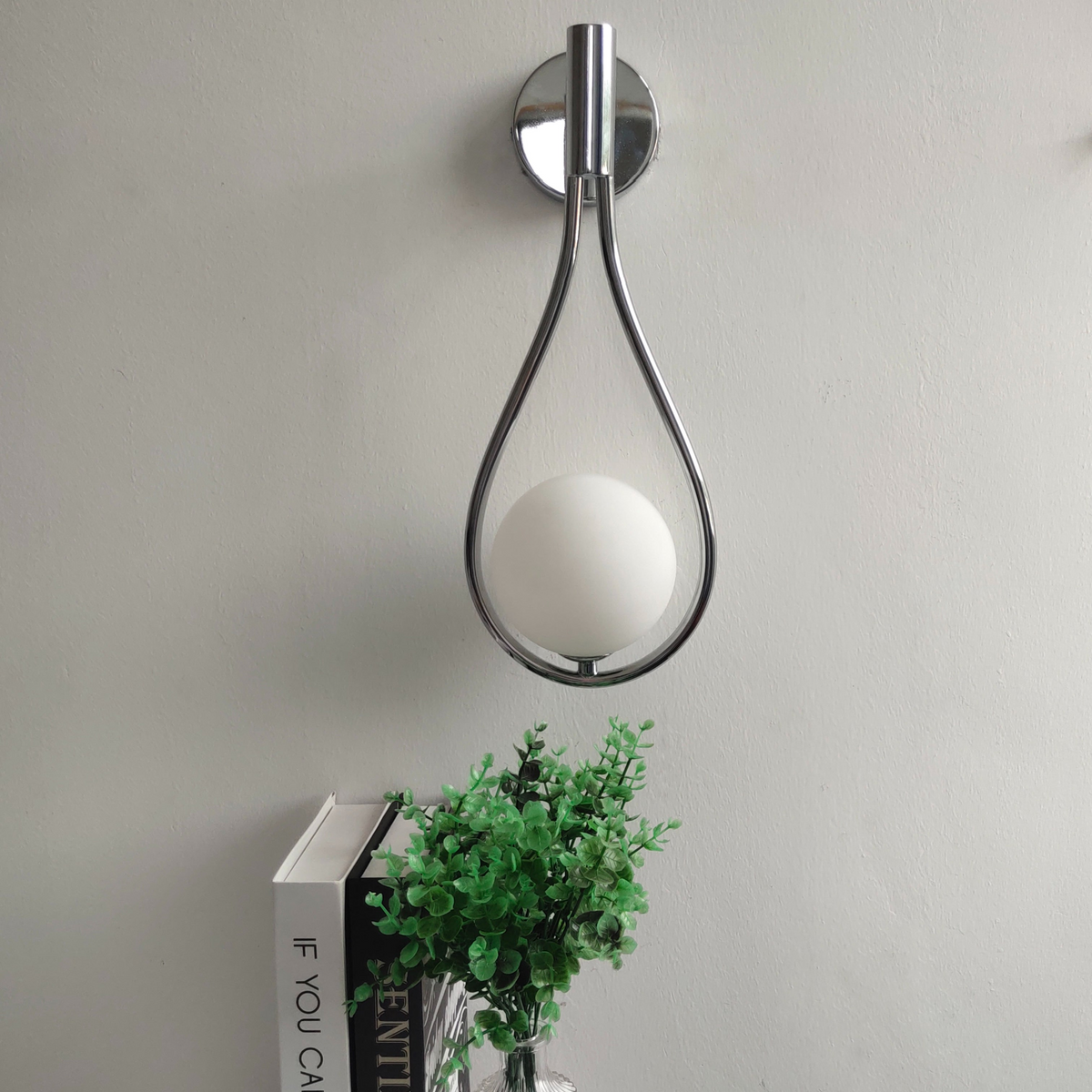 Silver teardrop globe wall sconce on a grey wall above books and a plant