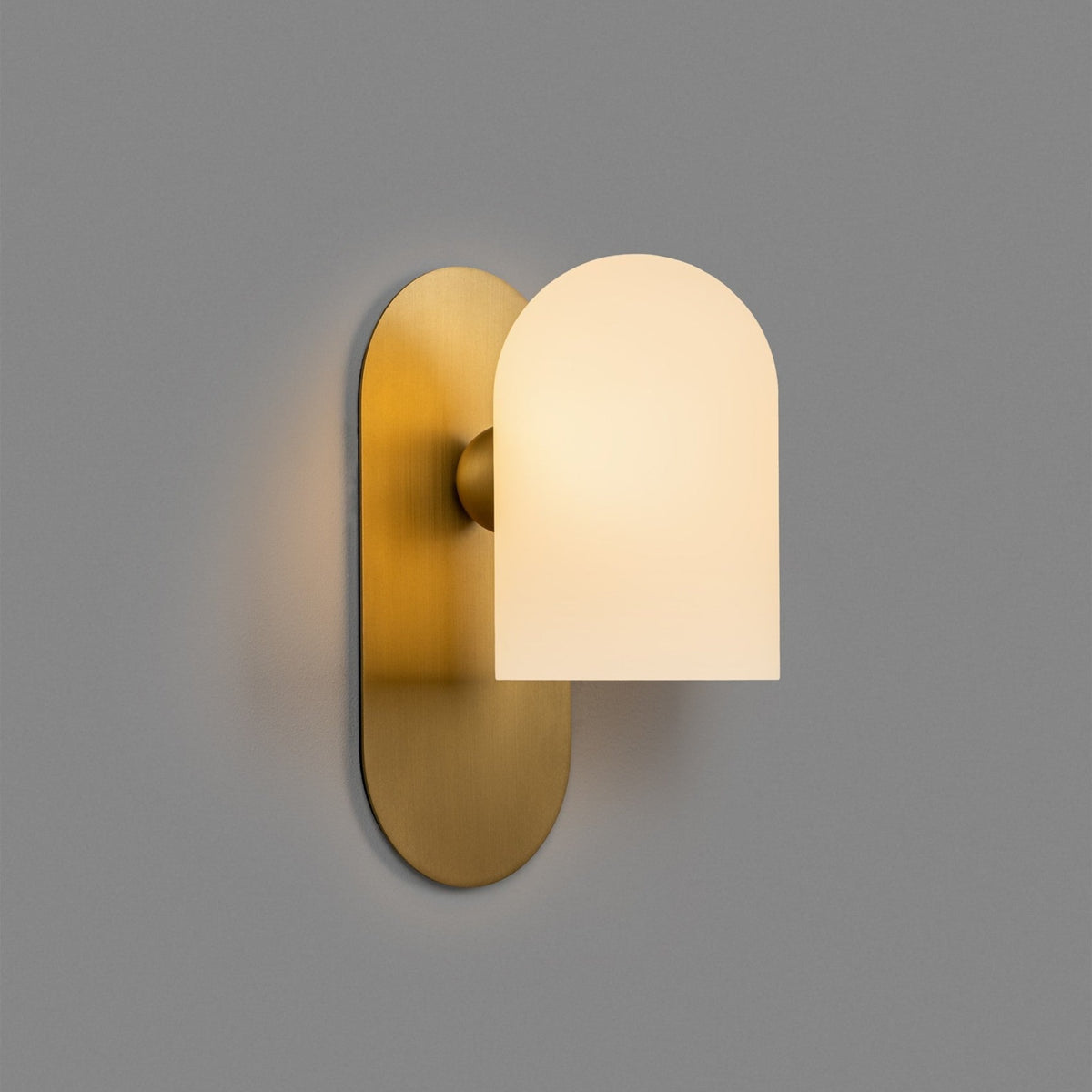 Capsula Opalina brass wall sconce glowing warm on a grey wall