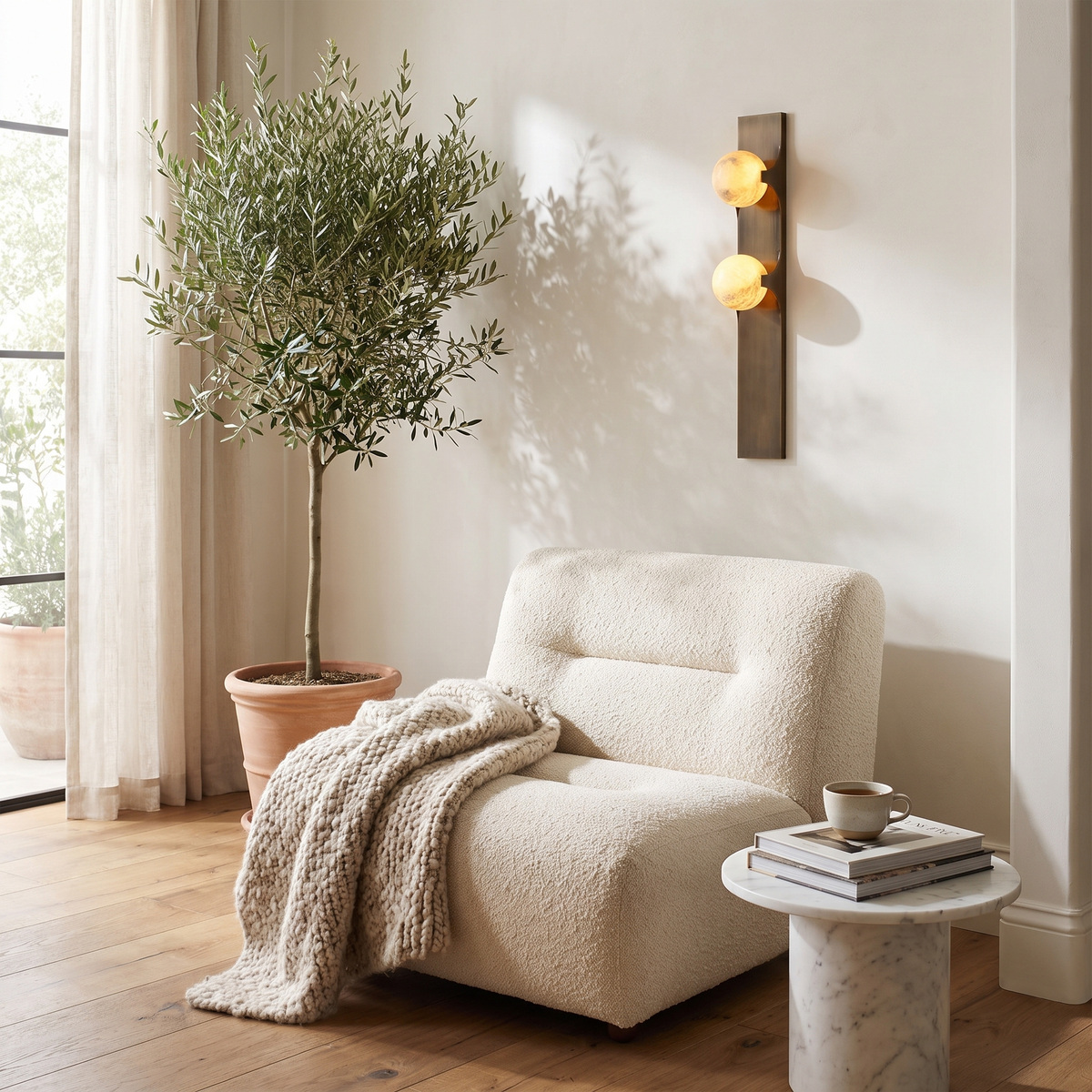 Alabaster wall sconce above a bouclé lounge chair in a bright reading nook with an olive tree