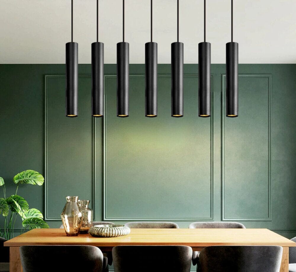 Row of black Flauto tube pendants over a dining table against green panelling